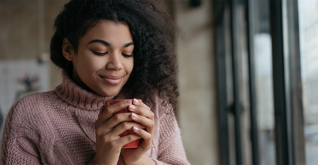 Woman breathing in herbal tea, eyes closed, natural hair, pink sweater, indoors, calm, relaxed, content